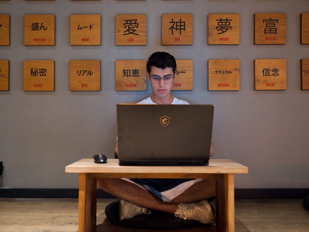 Joven estudiando japonés con letras japonesas de fondo Man sitting cross-legged using laptop, surrounded by Japanese text tiles.