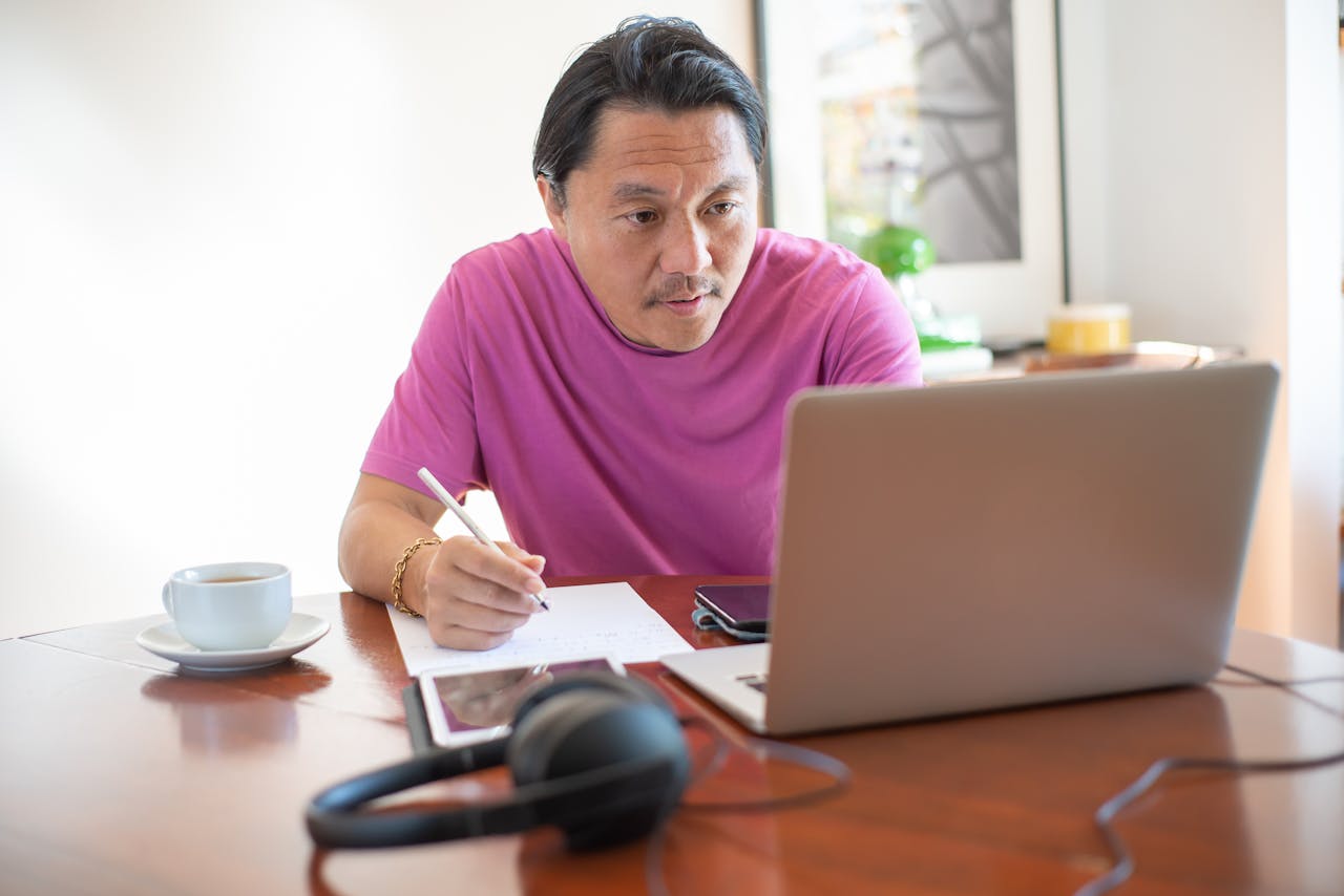 Services Asian man in pink shirt using laptop and pen at home office in Portugal.